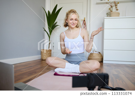 Portrait of beautiful fitness blogger, recording video on digital camera, showing workout exercises, explaining fitness movements or yoga training to followers, sits on rubber mat 113331074