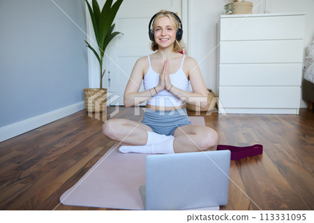Portrait of young, relaxed woman in headphones, listens to meditation music on laptop, practice yoga on rubber mat, holding hands clasped together in namaste sign Portrait of young, relaxed woman in headphones, listens to meditation music on laptop, practice yoga on rubber mat, holding hands clasped together in namaste sign 113331095