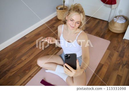 Portrait of woman having live stream training session, using her smartphone to create workout content, blogging during exercises or doing yoga, sitting on rubber mat at home Portrait of woman having live stream training session, using her smartphone to create workout content, blogging during exercises or doing yoga, sitting on rubber mat at home 113331098