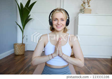 Portrait of blond young woman meditating, practise yoga, using yoga podcast to workout at home, sits on rubber mat 113331106
