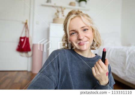 Portrait of young makeup artist, beauty blogger showing new lipstick, recording video in her room, smiling and expressing positivity Portrait of young makeup artist, beauty blogger showing new lipstick, recording video in her room, smiling and expressing positivity 113331110
