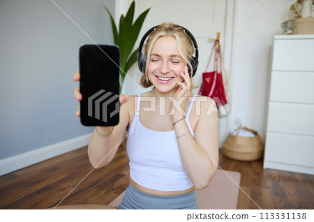 Portrait of smiling, beautiful young woman, showing her smartphone screen, wearing headphones, looking satisfied with sound quality 113331138