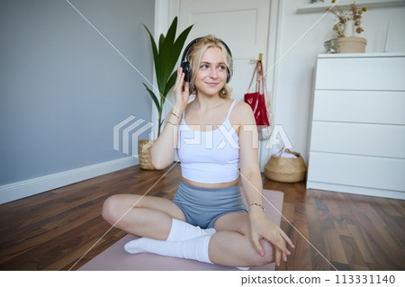 Portrait of happy, beautiful athletic woman, working out at home, listening music to boost energy while active training session, using wireless headphones and rubber yoga mat 113331140