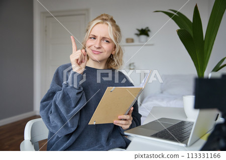 Portrait of young creative woman, content maker, sitting in room, working from home, using laptop, holding notebook, raising finger, has an idea, eureka gesture Portrait of young creative woman, content maker, sitting in room, working from home, using laptop, holding notebook, raising finger, has an idea, eureka gesture 113331166