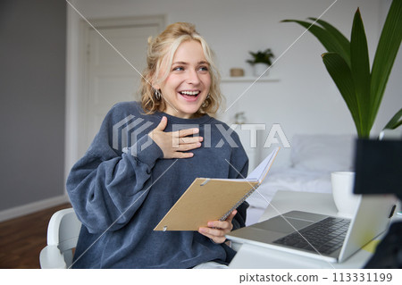 Portrait of smiling, beautiful young blond woman, student working on assignment from home, online learning in her bedroom, talking to video camera, chatting, holding notebook 113331199