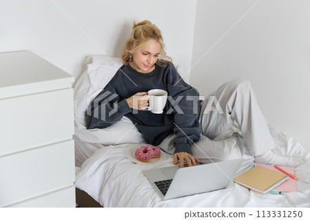 Portrait of young beautiful blond woman, feeling lazy at home, lying in bed with cup of tea, doughnut and laptop, studying remotely or watching movie online 113331250