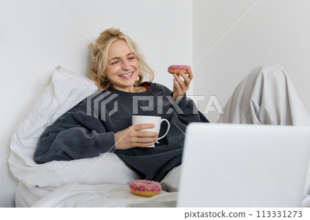 Lifestyle and people concept. Happy blond woman lying in bed with food, drinking tea and eating doughnut, resting at home, spending time alone, watching tv show 113331273