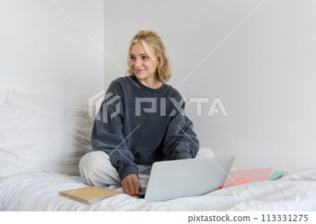 Portrait of happy blond woman, freelancer working from home, sitting on bed with laptop and notebooks. Student doing homework in bedroom, connects to online class via video chat 113331275