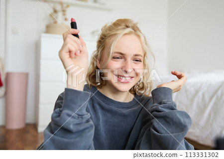 Portrait of young social media influencer, woman recording a video with beauty products, showing her makeup on camera, holding lipstick and smiling, sitting on floor 113331302
