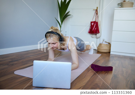 Portrait of young athletic woman in wireless headphones, standing in plank, following online workout video, using laptop to join training session on the internet Portrait of young athletic woman in wireless headphones, standing in plank, following online workout video, using laptop to join training session on the internet 113331360