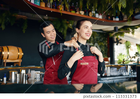Friendly bartender helping coworker to adjust apron strap Friendly bartender helping coworker to adjust apron strap 113331373