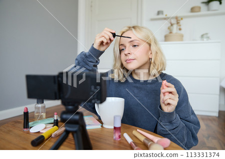 Portrait of blond smiling woman records a lifestyle blog, vlogger or makeup artist recording video for social media, holding mascara, reviewing beauty products for followers online Portrait of blond smiling woman records a lifestyle blog, vlogger or makeup artist recording video for social media, holding mascara, reviewing beauty products for followers online 113331374