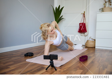 Portrait of young woman, fitness instructor recording video of her standing in a side plank, doing workout at home on camera, creating content for social media Portrait of young woman, fitness instructor recording video of her standing in a side plank, doing workout at home on camera, creating content for social media 113331481