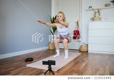 Portrait of woman, fitness instructor at home, recording video about workout, showing how to do leg exercises, squats with elastic resistance band, working out indoors on yoga mat 113331493