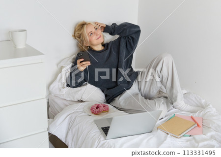 Lifestyle and weekend concept. Young smiling woman, relaxing at home, lying in bed with cup of tea and doughnut, using laptop and smartphone in bedroom 113331495
