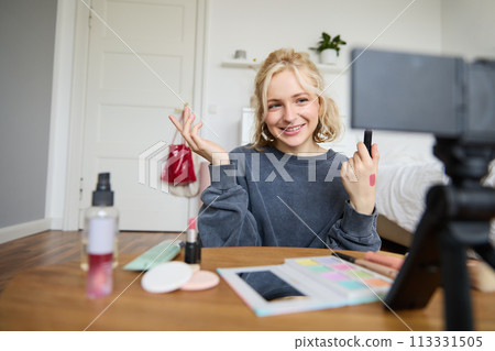Image of young stylish woman, blogger recording a beauty lifestyle video of her picking best lipstick, showing lip balm swatches on her skin, sitting in front of digital camera in empty room Image of young stylish woman, blogger recording a beauty lifestyle video of her picking best lipstick, showing lip balm swatches on her skin, sitting in front of digital camera in empty room 113331505