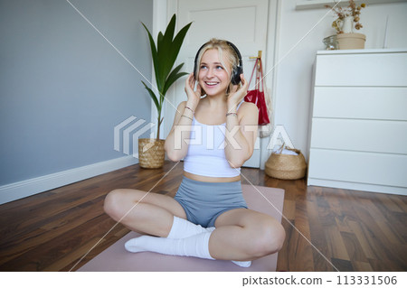 Young and sporty woman working out at home, doing fitness training alone on yoga mat, listens to music in wireless headphones Young and sporty woman working out at home, doing fitness training alone on yoga mat, listens to music in wireless headphones 113331506