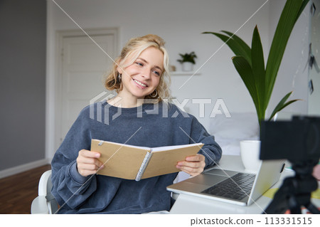 Portrait of smiling blond woman, sitting in bedroom, using laptop and digital camera, recording video for lifestyle blog, reading, using her notebook Portrait of smiling blond woman, sitting in bedroom, using laptop and digital camera, recording video for lifestyle blog, reading, using her notebook 113331515