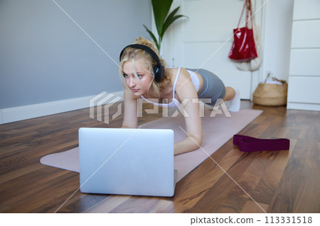 Portrait of young sporty woman doing workout, looking at fitness video on laptop in wireless headphones, standing in plank on yoga mat, following exercise tutorial 113331518