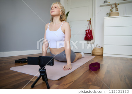 Portrait of young blogger, yoga content creator, showing exercises, recording video of herself working out at home on rubber mat 113331523