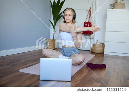 Portrait of young fitness woman doing warm-up exercises, looking at workout video on laptop, stretching arms, wearing headphones 113331524