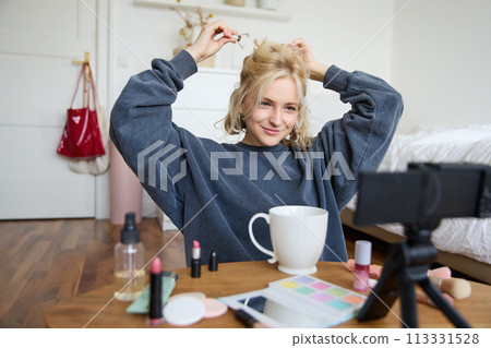 Lifestyle blogger records video of herself while doing hairstyle, showing makeup lifehacks for social media followers, vlogging, using camera for her blog, sitting in a room, drinking tea Lifestyle blogger records video of herself while doing hairstyle, showing makeup lifehacks for social media followers, vlogging, using camera for her blog, sitting in a room, drinking tea 113331528