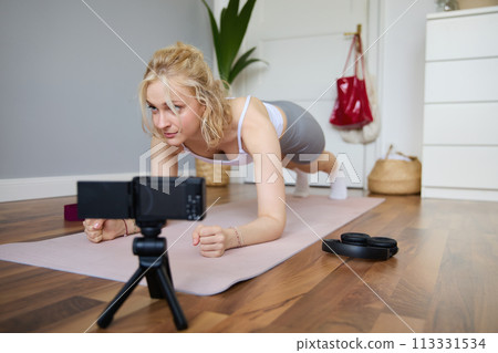 Young woman, personal fitness instructor records video of herself standing in a plank, using rubber yoga mat and digital camera at home 113331534
