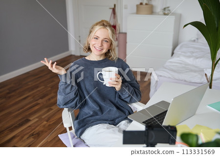 Image of young woman, social media influencer, editing her video on laptop, sits in a room with computer and digital camera, drinking coffee, smiling at camera Image of young woman, social media influencer, editing her video on laptop, sits in a room with computer and digital camera, drinking coffee, smiling at camera 113331569