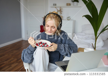 Portrait of smiling young blond woman in headphones, sitting in room, watching movie on laptop, eating breakfast and drinking tea, having lunch in front of computer screen 113331585