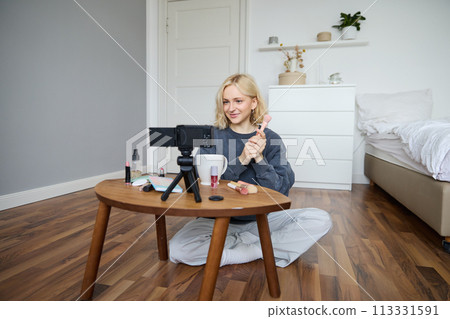 Portrait of beautiful smiling woman, recording video in her room, has camera on coffee table, reviewing makeup, doing lifestyle vlog for social media account, records a tutorial Portrait of beautiful smiling woman, recording video in her room, has camera on coffee table, reviewing makeup, doing lifestyle vlog for social media account, records a tutorial 113331591