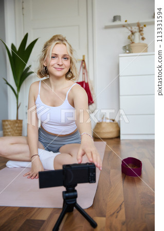 Vertical shot of woman sitting on yoga mat, recording video on digital camera, vlogging about workout at home and fitness lifestyle, showing how to stay fit and healthy indoors Vertical shot of woman sitting on yoga mat, recording video on digital camera, vlogging about workout at home and fitness lifestyle, showing how to stay fit and healthy indoors 113331594