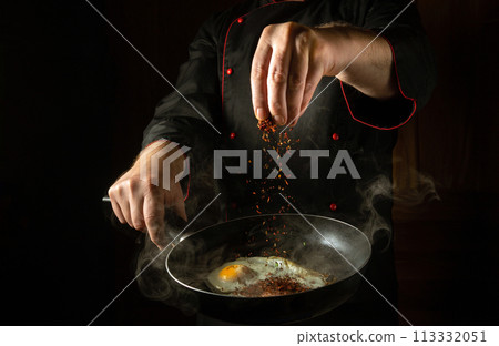 The cook adds aromatic spices to a steaming hot frying pan with fried eggs. Low key concept for a breakfast menu for a hotel with advertising space. Grande cuisine The cook adds aromatic spices to a steaming hot frying pan with fried eggs. Low key concept for a breakfast menu for a hotel with advertising space. Grande cuisine 113332051