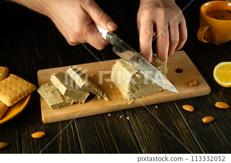 Cutting halva with a knife in the hands of a cook on a cutting board in a hotel kitchen. Low key concept of serving dessert with halva and cookies Cutting halva with a knife in the hands of a cook on a cutting board in a hotel kitchen. Low key concept of serving dessert with halva and cookies 113332052