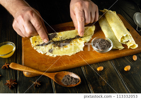 Making delicious pancakes. Chef hands roll a pancake with poppy seeds and honey on the kitchen table in a pub Making delicious pancakes. Chef hands roll a pancake with poppy seeds and honey on the kitchen table in a pub 113332053