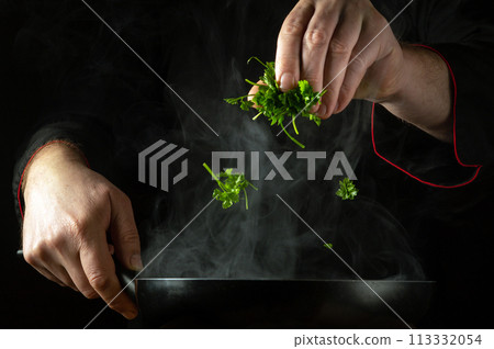 The chef adds fresh parsley to the hot frying pan by hand. Low key concept of preparing a delicious lunch in a restaurant kitchen. Grande cuisine The chef adds fresh parsley to the hot frying pan by hand. Low key concept of preparing a delicious lunch in a restaurant kitchen. Grande cuisine 113332054