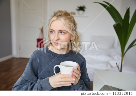 Close up portrait of smiling young blond woman, sits with cup of tea in bedroom, rests at home, enjoys her coffee 113332915