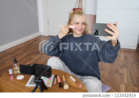 Portrait of beautiful blond girl doing makeup on camera, recording vlog using digital camera and live stream on mobile phone app, chatting with audience Portrait of beautiful blond girl doing makeup on camera, recording vlog using digital camera and live stream on mobile phone app, chatting with audience 113332919