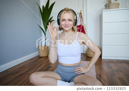 Portrait of young smiling beautiful woman, wearing wireless headphones, working out at home, sitting on rubber yoga mat, showing okay sign, satisfied with exercises Portrait of young smiling beautiful woman, wearing wireless headphones, working out at home, sitting on rubber yoga mat, showing okay sign, satisfied with exercises 113332925