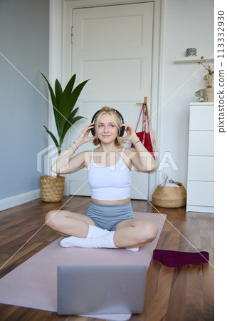 Vertical shot of smiling young woman using video tutorials for workout at home, sitting with laptop in wireless headphones on yoga mat, follows fitness instructor on social media, repeats exercises Vertical shot of smiling young woman using video tutorials for workout at home, sitting with laptop in wireless headphones on yoga mat, follows fitness instructor on social media, repeats exercises 113332930