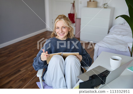Portrait of young woman reading her notes on journal, sitting at home on chair, smiling and looking happy, doing homework 113332951