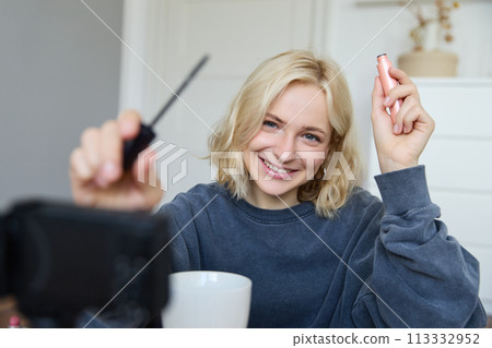 Portrait of young smiling woman in her room, recording video on camera, lifestyle vlog for social media, holding mascara, reviewing her makeup beauty products, showing how to use cosmetics 113332952