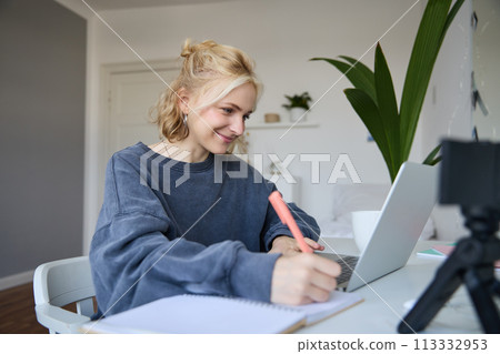 Portrait of smiling blond woman writing in notebook, making notes, recording content for social medial on digital camera, looking at laptop, working or studying 113332953