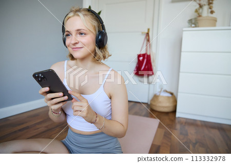 Portrait of young blond woman in headphones, turning on yoga, workout app on smartphone, choosing music on mobile phone application, sitting on rubber mat in room 113332978