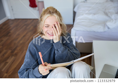 Portrait of charismatic blond girl, smiling woman in bedroom, holding notebook and pen, writing in journal or diary, creates to do list in planner 113332979