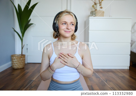 Portrait of young blond woman in headphones, feeling calm and relaxed after meditation, yoga training, sitting on yoga mat in her room 113332980