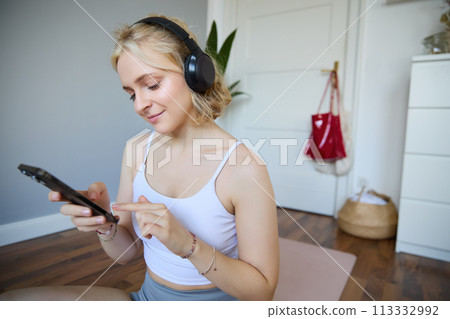 Portrait of young sporty woman in wireless headphones, sitting with smartphone on rubber mat, using workout training app, fitness application on her phone, listening in earphones 113332992