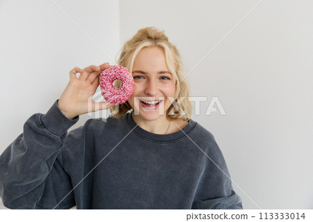 Close up portrait of cute young blond woman, showing pink doughnut near her face, smiling and looking happy 113333014