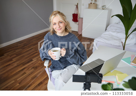 Image of young woman, social media influencer, editing her video on laptop, sits in a room with computer and digital camera, drinking coffee, smiling at camera 113333058