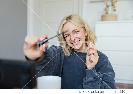 Portrait of young smiling woman in her room, recording video on camera, lifestyle vlog for social media, holding mascara, reviewing her makeup beauty products, showing how to use cosmetics 113333063