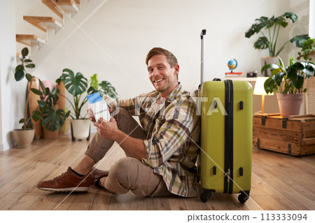 Portrait of male model with suitcase, sitting on floor, holding flight tickets and passport, going abroad, waiting for a taxi to airport 113333094